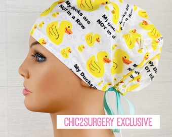 May include: A white scrub cap featuring a repeating pattern of yellow rubber ducks and light blue bubbles. The text "My Ducks are NOT in a Row" is printed in black across the fabric. The cap includes light blue ribbon ties at the back.