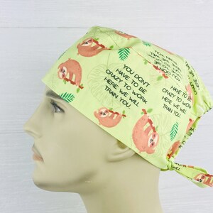 May include: A light green scrub hat with a pattern of sloths hanging from branches. The hat has a tie at the back and the text "You don't have to be crazy to work here we will train you" is printed on the hat.