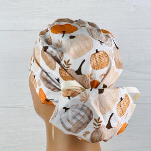 May include: A white scrub cap with a repeating pattern of pumpkins in shades of orange, brown, and beige. The cap has a matching bow and ties. The design is autumnal and festive.