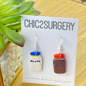 May include: A pair of dangle earrings featuring a white jar with a blue lid and a brown jar with an orange lid. The jars have black eyes and are on a white card with the text "CHIC2SURGERY" and "Handmade Medical Accessories".