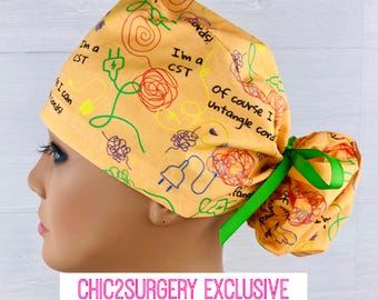 May include: A yellow surgical scrub hat with a green ribbon. The hat has a print of tangled cords and the text "I'm a CST, of course I untangle cords!"