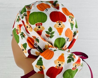 May include: A white scrub hat with a pattern of pumpkins, leaves, and dogs wearing hats. The hat has a burgundy tie.