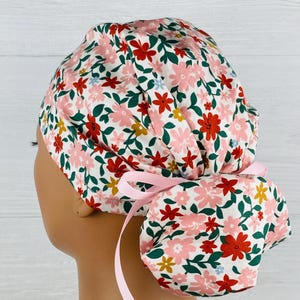 May include: A scrub cap featuring a white background with a floral pattern of pink, red, and yellow flowers with dark green leaves. The cap has a gathered back section for hair and is secured with a light pink ribbon tied in a bow at the nape.
