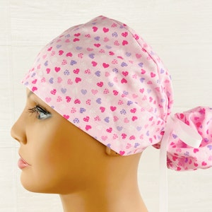 May include: Pink surgical scrub hat with a white background and a pattern of pink, purple, and white hearts. The hat has a ponytail opening and a white ribbon tie.