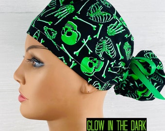 May include: A black scrub cap featuring a vibrant green skeleton bone and skull print. The cap includes a gathered back with a green ribbon tie and a rectangular graphic at the bottom right corner with the text GLOW IN THE DARK.