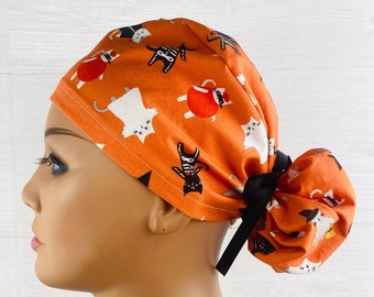 May include: Orange scrub hat with a black and white cat print. The hat has a black tie that can be tied in a bun at the back.