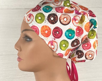 May include: A scrub cap featuring a white background with a colorful pattern of various donuts in shades of pink, orange, green, blue, and brown. The cap has a gathered back and is secured with two bright pink ribbons tied at the nape of the neck.