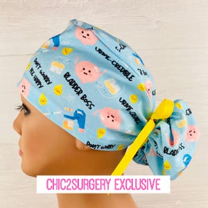 May include: A blue and pink patterned surgical scrub hat with yellow ties. The hat features a cartoon bladder with the text "Bladder Boss" and "Urine-Credible".