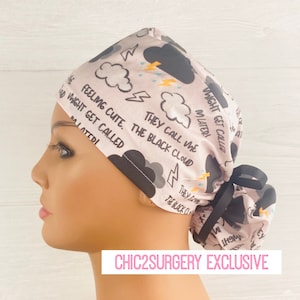 May include: A light pink surgical scrub cap with a black cloud and lightning bolt pattern. The cap features the text "Feeling Cute. The Black Cloud Might Get Called Later!" and a black ribbon tie.