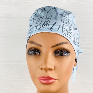 Puede incluir: Un gorro quir&uacute;rgico azul claro con un patr&oacute;n repetido de instrumentos m&eacute;dicos y la palabra "NEURO" en un cuadrado con un s&iacute;mbolo de coraz&oacute;n. El gorro tiene lazos en la parte posterior y est&aacute; hecho de una tela suave.