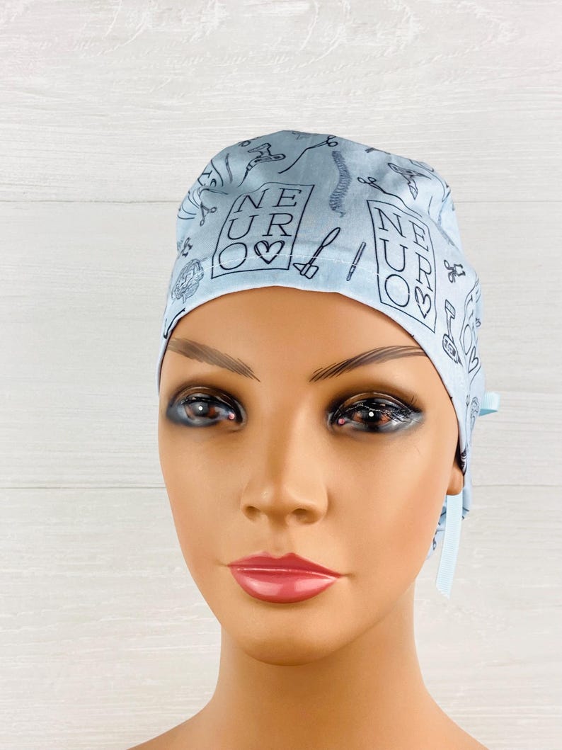 Puede incluir: Un gorro quir&uacute;rgico azul claro con un patr&oacute;n repetido de instrumentos m&eacute;dicos y la palabra "NEURO" en un cuadrado con un s&iacute;mbolo de coraz&oacute;n. El gorro tiene lazos en la parte posterior y est&aacute; hecho de una tela suave.