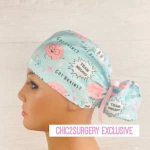 Pu&ograve; includere: Un berretto chirurgico azzurro chiaro con un design a cervello di cartone animato rosa e bianco. Il berretto ha una cravatta rosa e il testo "Got Brains?" e "Team Neuro" stampato su di esso. Chic2Surgery Exclusive.
