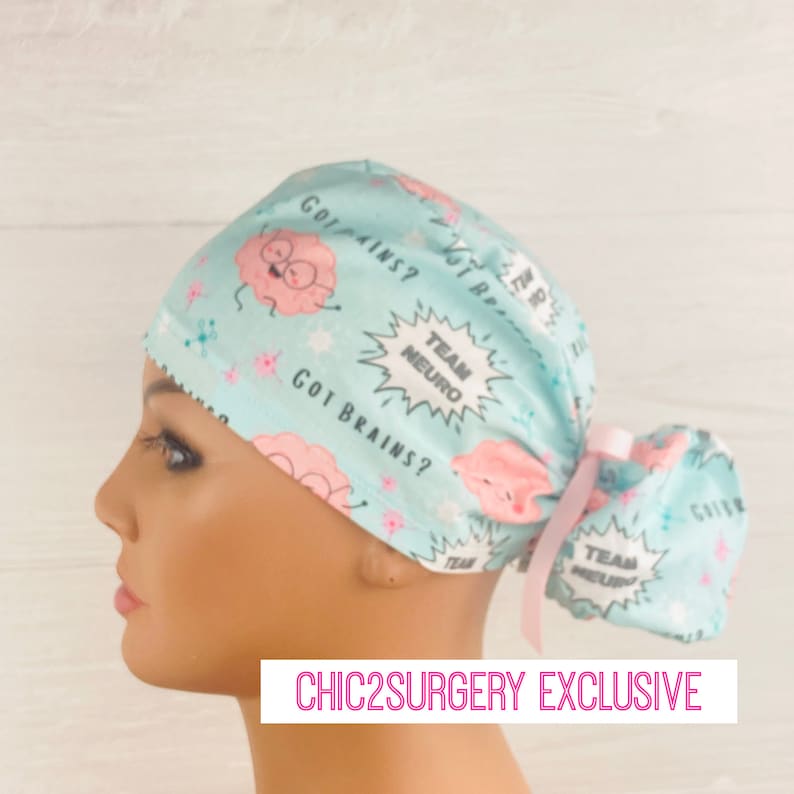 Pu&ograve; includere: Un berretto chirurgico azzurro chiaro con un design a cervello di cartone animato rosa e bianco. Il berretto ha una cravatta rosa e il testo "Got Brains?" e "Team Neuro" stampato su di esso. Chic2Surgery Exclusive.