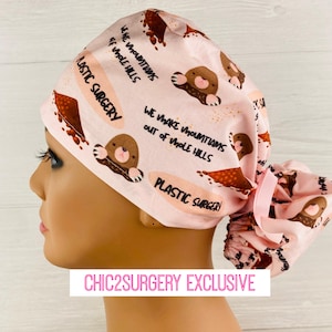 May include: A light pink scrub cap featuring a repeating pattern of brown moles, piles of dirt, and the text "We make mountains out of mole hills" and "Plastic Surgery". The cap includes a gathered back section with a tied pink ribbon closure.