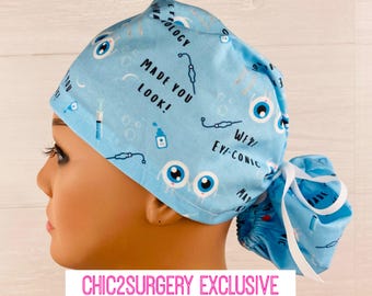 May include: Blue surgical scrub hat with a white tie. The hat has a pattern of eyes, syringes, and the text "Made you look!" and "We're eye-conic".