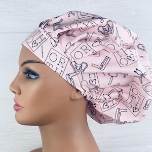May include: A light pink scrub cap featuring a repeating black line art pattern of orthopedic surgical tools, bones, and the text ORTHO with a heart symbol. The cap has a gathered, pleated design with a rounded crown and a fitted band around the head.