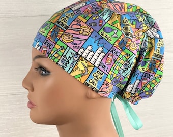 May include: A scrub cap featuring a colorful comic-style print with medical illustrations of anesthesia equipment, vials, and syringes. The fabric includes text such as Keep Calm and Call Anesthesia, Propofol, and ROC. It has a mint green ribbon tie.