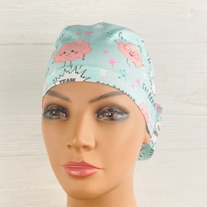 Pu&ograve; includere: Un berretto chirurgico azzurro chiaro con un design a cartone animato di un cervello rosa. Il berretto ha il testo "Got Brains" e "Team Brain Power".
