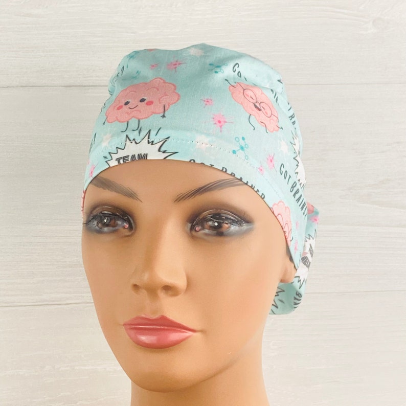 Pu&ograve; includere: Un berretto chirurgico azzurro chiaro con un design a cartone animato di un cervello rosa. Il berretto ha il testo "Got Brains" e "Team Brain Power".