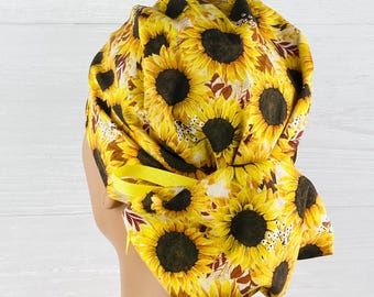 May include: A yellow sunflower patterned scrub cap with a yellow ribbon. The cap features a repeating pattern of sunflowers with brown centers and yellow petals. The background is white.