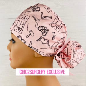 May include: A light pink scrub cap featuring a repeating black line art pattern of orthopedic medical symbols, bones, and the text ORTHO with a heart. The cap includes a gathered back section secured with a light pink ribbon tie.