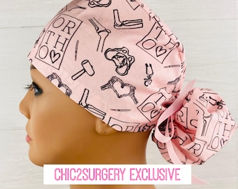 May include: Pink surgical scrub hat with a black and white print of orthopedic medical symbols and the text "ORTHO" with a heart. The hat has a pink tie.