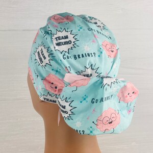 Pu&ograve; includere: Un berretto chirurgico blu chiaro con un laccio rosa. Il berretto ha un motivo di cervelli dei cartoni animati con il testo "Team Neuro" e "Got Brains?" ripetuto sul tessuto.