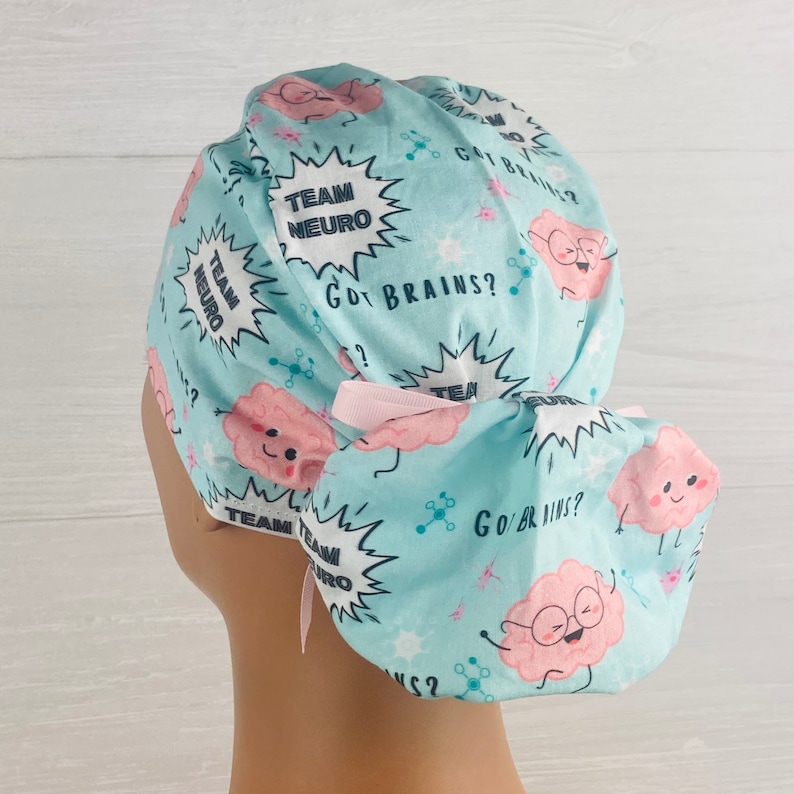 Pu&ograve; includere: Un berretto chirurgico blu chiaro con un laccio rosa. Il berretto ha un motivo di cervelli dei cartoni animati con il testo "Team Neuro" e "Got Brains?" ripetuto sul tessuto.