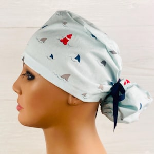 May include: A light blue scrub cap featuring a repeating pattern of small grey and dark blue shark fins, white wave accents, and red buoys. The cap has a gathered back section secured with a dark blue ribbon tie, creating a ponytail pouch style.