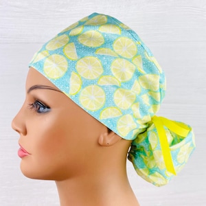 May include: A light blue and yellow scrub cap featuring a pattern of lemon slices. The cap is tied in a ponytail style with a yellow ribbon. The background is a light blue color.
