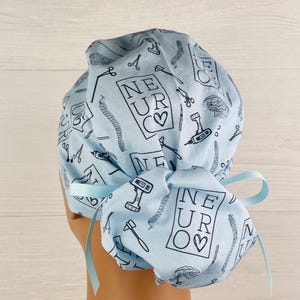 Puede incluir: Gorro quir&uacute;rgico azul claro con un patr&oacute;n repetido de herramientas m&eacute;dicas y el texto "NEUR♡". El gorro tiene una cinta en la parte posterior. El dise&ntilde;o incluye tijeras, taladros e ilustraciones de cerebro.