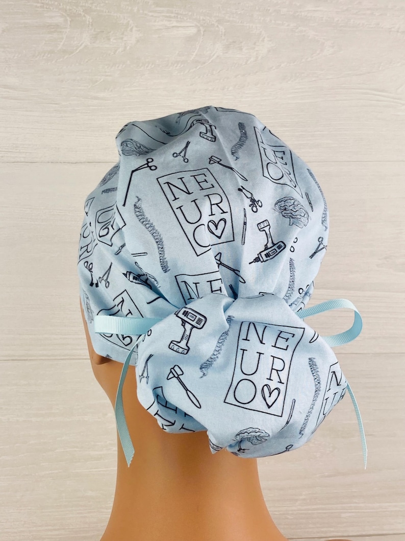 Puede incluir: Gorro quir&uacute;rgico azul claro con un patr&oacute;n repetido de herramientas m&eacute;dicas y el texto "NEUR♡". El gorro tiene una cinta en la parte posterior. El dise&ntilde;o incluye tijeras, taladros e ilustraciones de cerebro.