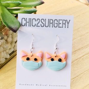 May include: A pair of dangle earrings featuring a peach-colored cat face with a light blue medical mask. The earrings are on a white card with the text "CHIC2SURGERY" and "Handmade Medical Accessories".