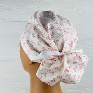 May include: A white scrub cap with a repeating pattern of pink bows. The cap is tied in the back with a white ribbon. The fabric is a light cotton, and the overall design is feminine and delicate.