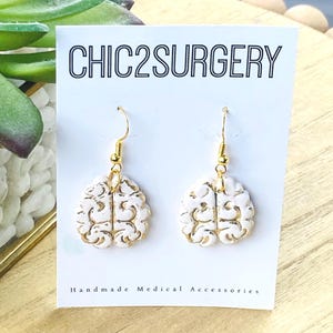 May include: White brain-shaped earrings with gold detailing on a white card with the text "CHIC2SURGERY" and "Handmade Medical Accessories".