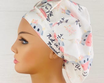 May include: A white scrub cap featuring a repeating pattern of cartoon cow faces with pink noses, small floral clusters, and the black cursive text Not in the Mooood. The fabric cap has a gathered design with a wide front band and a pleated crown.