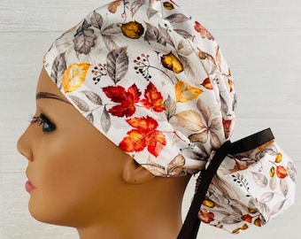 May include: A scrub cap featuring a white background with a pattern of autumn leaves in shades of red, orange, yellow, and grey. The cap has a gathered back section secured with a solid black ribbon tied into a bow, providing a snug fit.