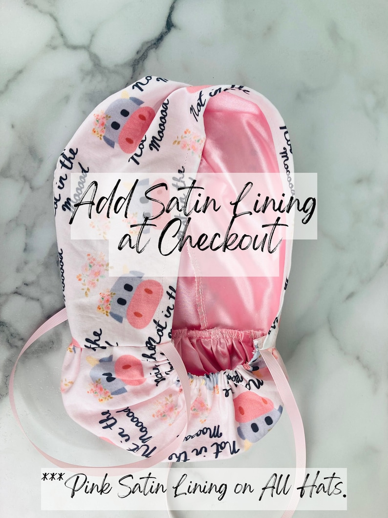 Puede incluir: Un gorro rosa y blanco con un estampado de vaca en el exterior y un forro de sat&eacute;n rosa. El gorro tiene un cord&oacute;n y el texto "Not in the mood" y "Mooooa" est&aacute; impreso en el exterior. El texto "Add Satin Lining at Checkout" tambi&eacute;n es visible.