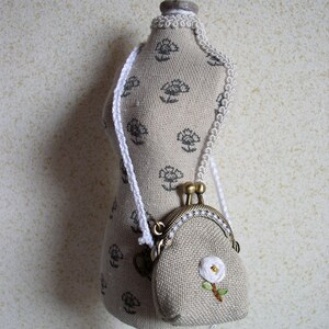 Peut inclure: Un petit sac à main en lin beige avec une fleur blanche brodée sur le devant. Le sac à main a une fermeture en laiton et une bandoulière en cordon blanc. Le sac à main est présenté sur un mannequin en tissu blanc.