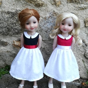 May include: Two dolls wearing white dresses with red and navy blue sashes and white collars. The dolls have blonde and brown hair and are wearing white shoes.