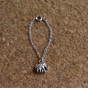 Peut inclure: Un collier avec un pendentif en forme d'éléphant argenté. Le pendentif, orné d'un motif floral sur le côté, est suspendu à une chaîne délicate. Le collier est présenté sur un fond marron.