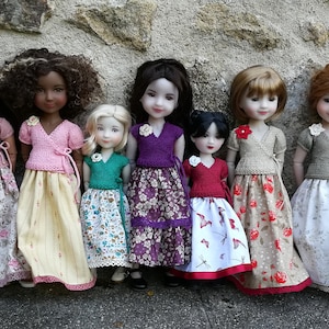 May include: Nine dolls wearing different outfits, standing in a row against a stone wall. The dolls are all wearing skirts and tops, and some have floral patterns.