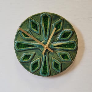 May include: A circular ceramic wall clock featuring a deep green glaze with a textured, crystalline finish. The clock face displays a radial, geometric pattern of recessed diamond and petal shapes. Three thin, gold-coloured metal hands are centred.