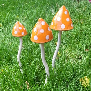 Może przedstawiać: Trzy ceramiczne grzyby z białymi kropkami na pomarańczowych kapeluszach. Grzyby są ułożone na trawiastym obszarze.