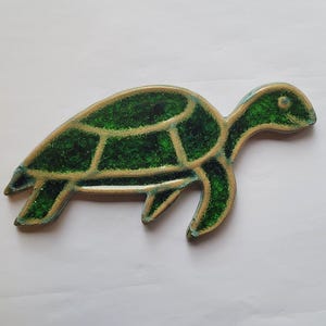 Könnte beinhalten: Dekorative Schildkrötenfigur mit grünem und goldenem Design. Die Schildkröte hat ein segmentiertes Schalenmuster mit goldener Kontur. Der Körper ist leuchtend grün mit glänzendem Finish. Die Figur befindet sich auf einer weißen Oberfläche.