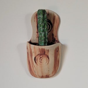 May include: A handmade, wall-mounted ceramic planter with a light brown, wood-grain finish. The planter holds a green cactus. The planter has a curved shape with a decorative spiral design. A unique piece for home decor.