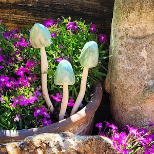 Op de afbeelding: Drie keramische paddenstoelsculpturen met lichtgroene doppen en witte stelen staan tussen levendige paarse bloemen en groen gebladerte in een terracotta pot. De paddenstoelen geven een grillige toets aan de tuin.