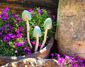 May include: Three ceramic mushroom sculptures with pale green caps and white stems are nestled among vibrant purple flowers and green foliage in a terracotta pot. The mushrooms add a whimsical touch to the garden setting.