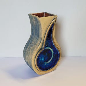 May include: A small, ceramic vase with a unique, abstract design. The vase is primarily yellow with blue and brown accents. The blue accents are in the shape of a swirl.