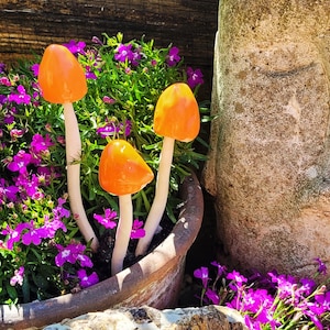 May include: A plant pot filled with purple flowers and several decorative mushrooms. The mushrooms have bright orange caps and white stems. A large, weathered stone structure is partially visible in the background.